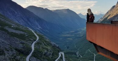 Vrouw met rugzak poseert in de punt van het uitkijkpunt boven Trollstigen. In de verte zie je bergtoppen en de kronkelende bergpas