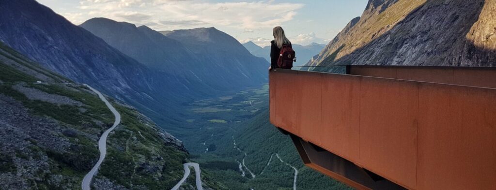 Vrouw met rugzak poseert in de punt van het uitkijkpunt boven Trollstigen. In de verte zie je bergtoppen en de kronkelende bergpas