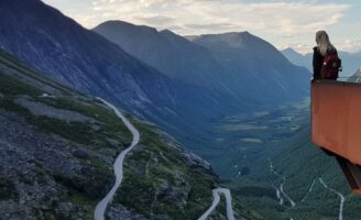 Vrouw met rugzak poseert in de punt van het uitkijkpunt boven Trollstigen. In de verte zie je bergtoppen en de kronkelende bergpas